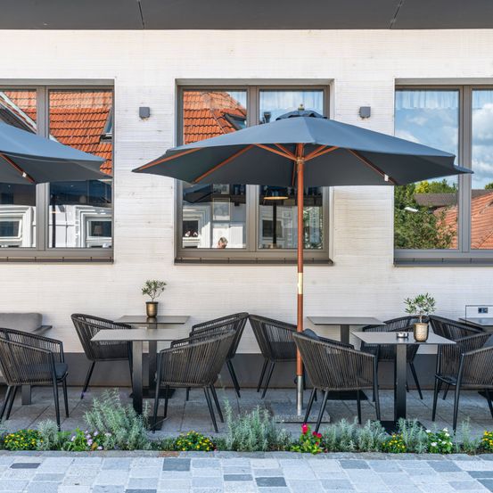 Eine Außensitzecke mit Tischen und Stühlen unter Schirmen vor einem Gebäude mit Glasfenstern und einem gefliesten Dach.