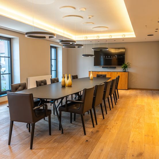 Ein leerer Konferenzraum mit einem langen Tisch, Stühlen und einem montierten Fernseher. Ein Heizkörper und eine Topfpflanze sind in der Nähe des Fensters.