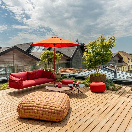 Eine Dachterrasse mit einem roten Sofa, einem runden Ottoman und einem Couchtisch unter einem orangefarbenen Schirm. Bäume und Pflanzen sind verstreut, mit Blick auf Häuser und einen bewölkten Himmel.