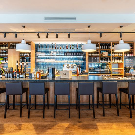 Ein Innenraumblick einer Bar mit Holzboden und Regalen voller Likörflaschen, einer Theke mit Barhockern und Pendelleuchten.