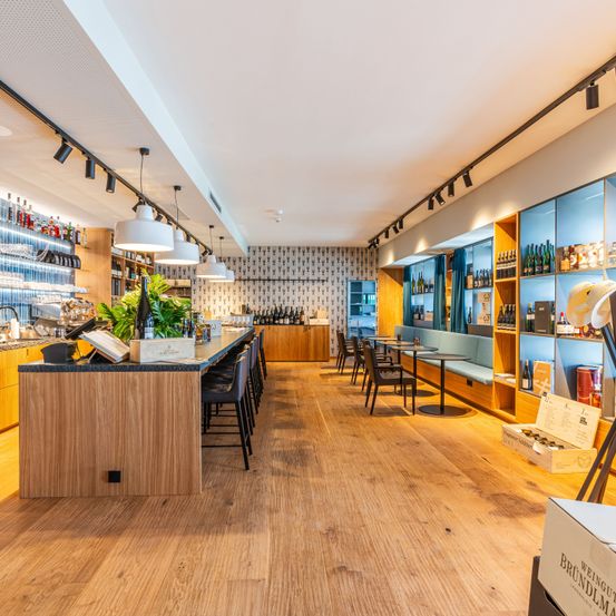 Eine moderne Bar mit Holzboden, weißen Wänden und Regalen voller Flaschen. Der Tresen hat Pflanzen und eine Speisekarte, mit Stühlen und Tischen in der Nähe.