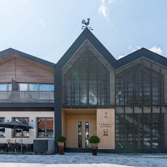 Die Vorderseite eines Hotels zeichnet sich durch ein modernes Design mit Holzelementen und Glasflächen aus. Es verfügt über einen Balkon mit Geländer und einen großen Schirm davor.