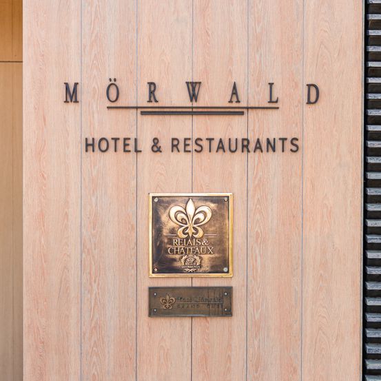 Eine hölzerne Tafel zeigt 'MORWALD HOTEL & RESTAURANTS' mit einer Fleur-de-Lis-Plakette darunter und einer kleineren Plakette mit französischem Text.
