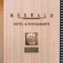 A wooden sign displays 'MORWALD HOTEL & RESTAURANTS' with a fleur-de-lis plaque below and a smaller plaque with French text.