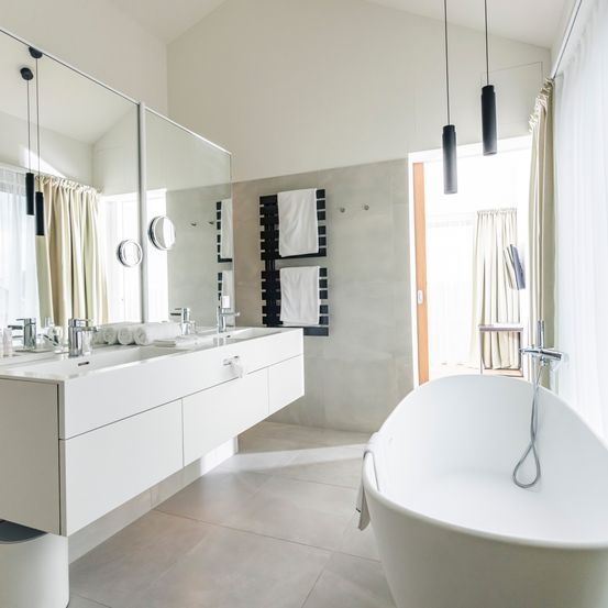 Modernes Badezimmer mit weißen Wänden, großem Spiegel, Doppelwaschbecken und Badewanne. Handtücher sind auf der Stange und der Boden ist gefliest.