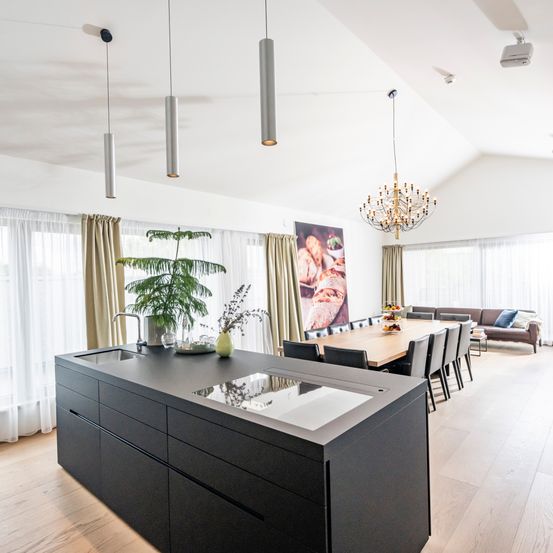 Eine moderne Küche mit einer großen Insel, dunklen Schränken und einer Kronleuchter. Ein Esstisch mit Stühlen, eine Couch und ein Gemälde im Hintergrund.