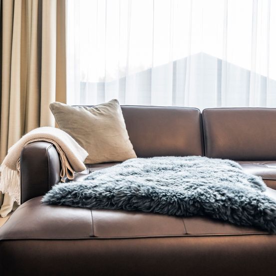 Ein modernes Wohnzimmer mit einem braunen Ledersofa und einer blauen Decke. Im Hintergrund sind ein weißer Vorhang und beige Vorhänge zu sehen.