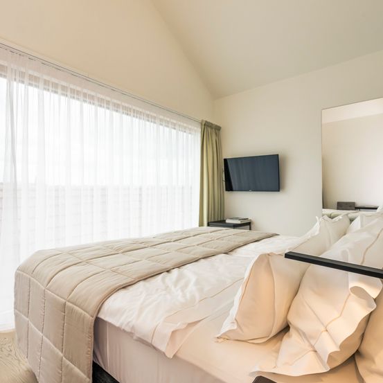 Ein geräumiges Schlafzimmer mit einem großen Bett, weißen Bettlaken, beigem Quilt und Kissen. Ein Flachbildfernseher und ein Spiegel sind an der Wand montiert. Ein Nachttisch steht neben dem Bett.