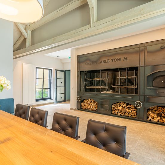 Ein Raum mit einem langen Holztisch und vier Lederstühlen vor einem großen Ofen mit einem Schild, das Küchentisch Toni M. lautet