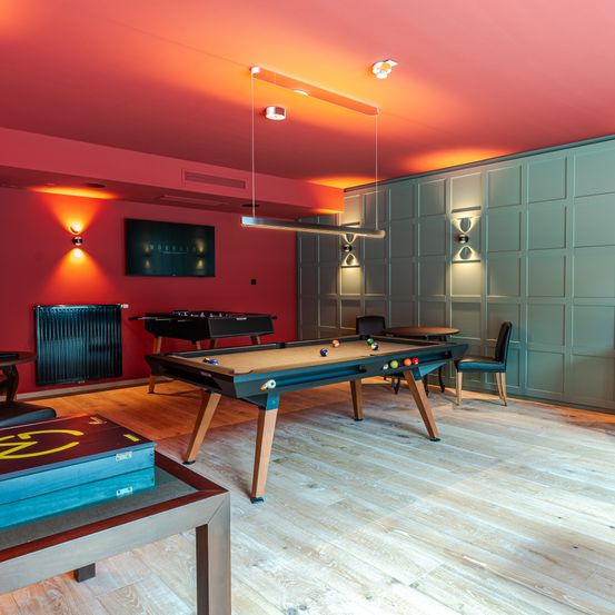 Ein Raum mit roten Wänden und Holzböden. Es gibt einen Billardtisch, Stühle und einen Heizkörper. Der Billardtisch hat Bälle und eine Lampe hängt von der Decke.
