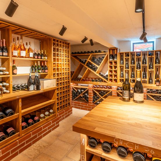 Ein gut organisierter Weinkeller mit Holzregalen und Ziegelwänden, der zahlreiche Weinflaschen enthält. Ein Holztisch im Vordergrund hat Flaschen darauf angeordnet, und die Decke hat ein paar Lampen.