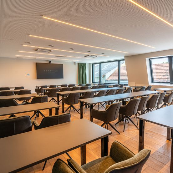 Ein gut beleuchteter Konferenzraum mit langen Tischen und Stühlen, einer großen Leinwand an der Wand und Fenstern mit Blick nach draußen.