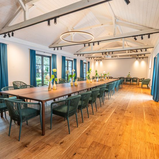 Ein leerer Essraum mit Holzboden, blauen Stühlen und einem langen Tisch mit Vasen und Blumen. Der Raum hat weiße Wände, Holzbalken und Fenster mit blauen Vorhängen.