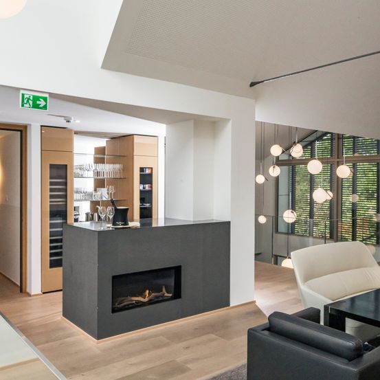 Ein modernes Interieur mit Kamin, Holzboden und Glaswand. Es gibt Sofas, einen Tisch und Weingläser auf einem Regal.