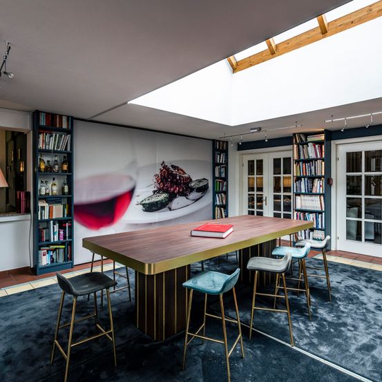 Ein Esszimmer mit einem langen Tisch, Weingläsern und einem Buch. Es gibt Stühle um den Tisch herum und eine Wand mit einem Gemälde eines Weinglases und Essen. Auf beiden Seiten des Raumes befinden sich Bücherregale mit Büchern.