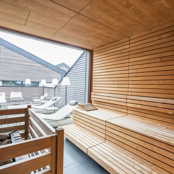 Ein moderner Holzsaunaraum mit einem großen Glasfenster, das den Blick nach draußen ermöglicht. Er hat Holzbänke, Lampen und Liegestühle.