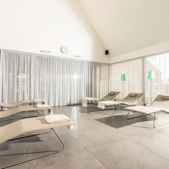 Ein Entspannungsraum mit mehreren Liegestühlen, einem weißen Vorhang und zwei grünen Stehlampen. Der Boden ist gefliest.