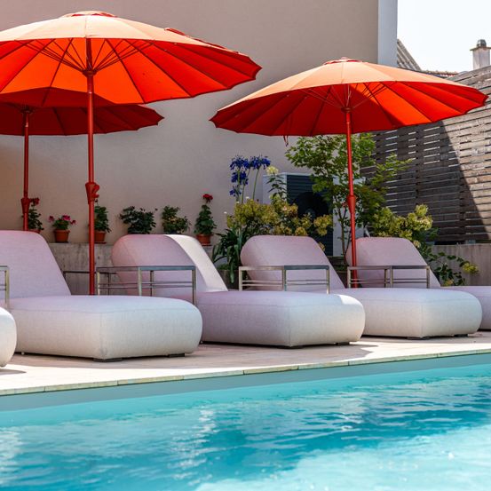 Mehrere Liegestühle stehen neben einem Pool mit roten Sonnenschirmen darüber. Der Pool hat klares Wasser, und es gibt Topfpflanzen und Blumen im Hintergrund.