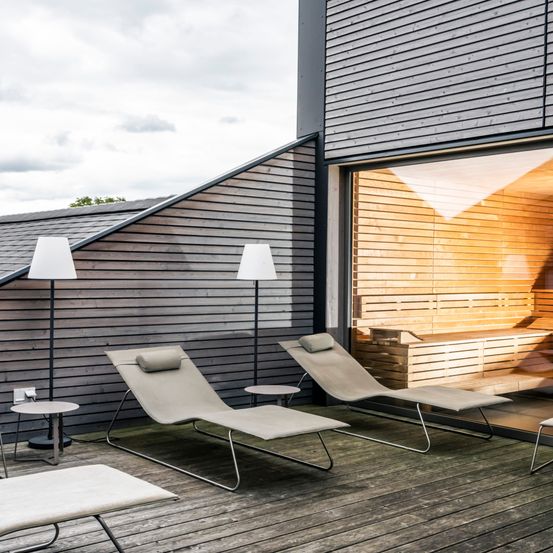 Eine Holzterrasse mit drei Liegestühlen, zwei mit Kissen, und zwei Stehlampen. Ein Saunaraum ist durch eine Glastür sichtbar.