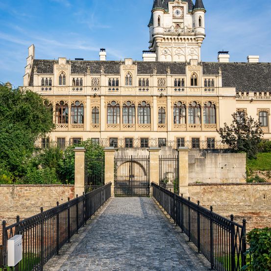 Ein großes Schloss mit vielen Fenstern und einem Turm mit einer Uhr steht auf einem Hügel. Ein Kopfsteinpflasterweg führt zu einem schwarzen Tor vor ihm.
