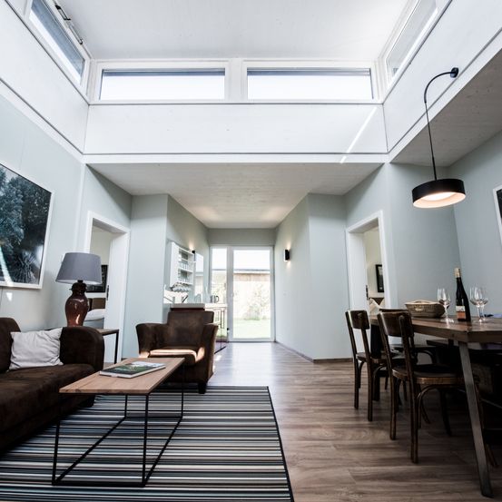 Ein modernes Wohnzimmer mit Holzboden, weißen Wänden und einem Oberlicht. Ein Sofa mit Kissen steht neben einem Couchtisch mit einem Buch. Ein Sessel und eine Lampe sind in der Nähe. Ein Esstisch mit Weingläsern und einer Flasche ist aufgestellt.