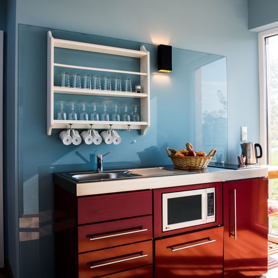 Eine kleine Küche mit blauen Wänden und roten Schränken. Sie verfügt über eine Mikrowelle, ein Waschbecken und einen Obstkorb auf der Arbeitsplatte. An der Wand befindet sich ein Regal mit Gläsern und Tassen.