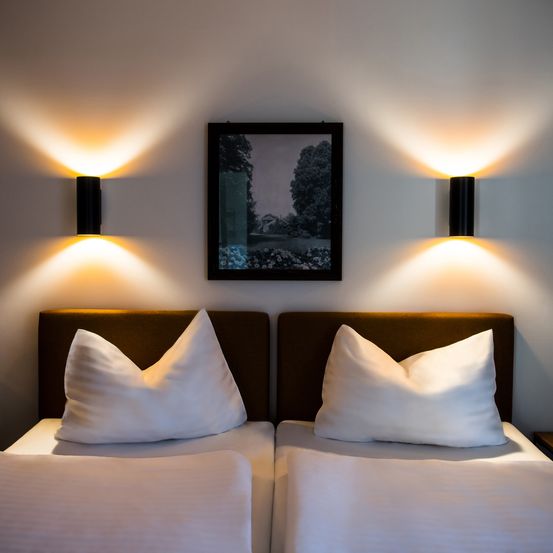 Ein Schlafzimmer mit zwei Betten, jedes mit einem weißen Kissen und braunem Kopfteil. Ein schwarz gerahmtes Bild hängt über den Betten. Zwei schwarze Wandleuchten beleuchten den Raum.