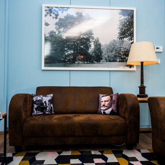 Ein braunes Sofa mit zwei Kissen, eines mit einem Schwarz-Weiß-Foto von Menschen, steht in einem Raum mit blauer Wand, einem großen gerahmten Bild und einer Lampe.