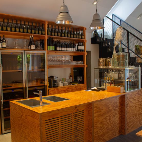 Eine Küche mit einer Holztheke hat zwei Waschbecken. Regale mit Flaschen, Weingläsern und Tassen an der Wand. Rechts ist eine Treppe.