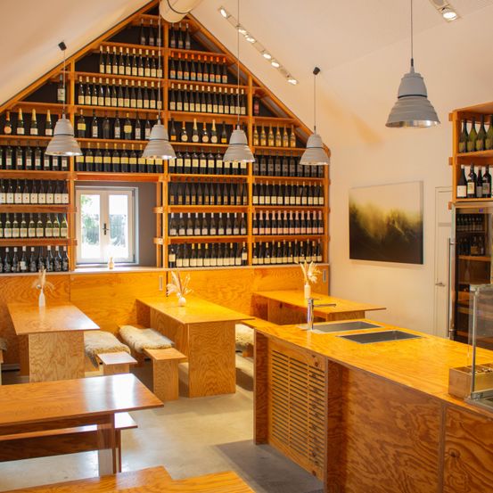 Ein Innenraum mit Holztischen, Bänken und einer dreieckigen Wand voller Weinflaschen. Es gibt einen Küchenbereich mit einem Doppelwaschbecken, und große Pendelleuchten hängen von der Decke.