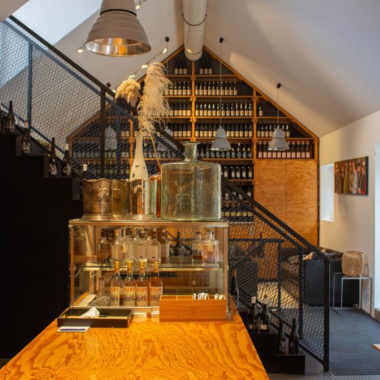 Ein Innenbereich einer Bar mit Holztheke und Regalen mit Likörflaschen. Ein Glaskasten mit mehreren Flaschen davor. Eine Treppe mit Geländern und hängenden Lampen in der Nähe.