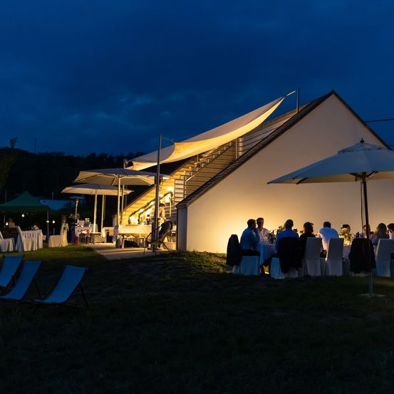 Eine nächtliche Zusammenkunft unter Schirmen und Lichtern bei einem Haus mit einer Treppe und Terrasse, mit sitzenden Gästen und einer Person in der Nähe der Treppe.