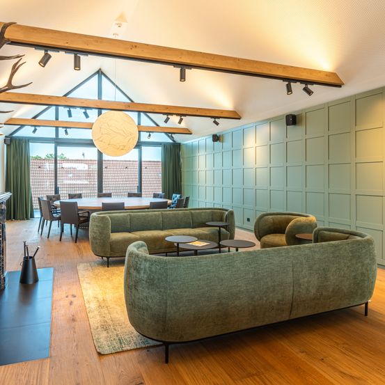 Ein modernes Interieur mit grünen Wänden und Holzboden, ausgestattet mit einem runden Tisch und Stühlen. Es gibt zwei grüne Sofas mit Tischen und einen Kamin auf der linken Seite. Eine Lampe hängt von der Decke.