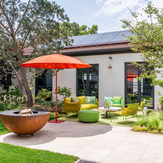Eine moderne Terrasse im Freien mit gelbem Sofa, grünen Stühlen und einem roten Sonnenschirm. Ein Feuerkorb und Solarmodule sind sichtbar.