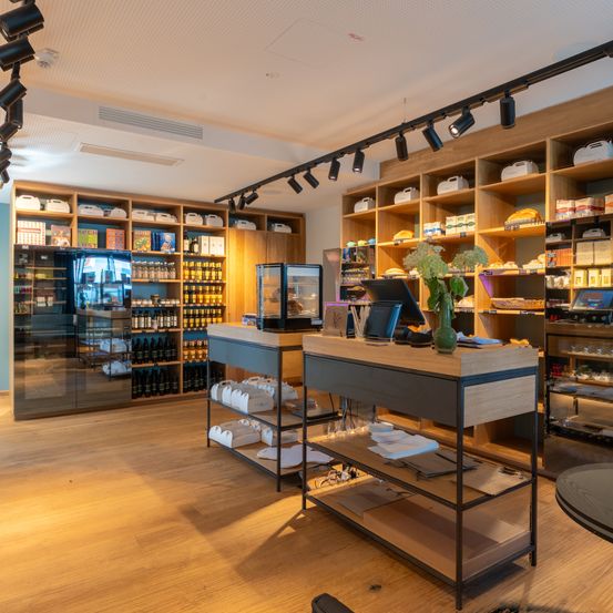 Das Innere eines Ladens mit Holzregalen gefüllt mit Artikeln und einer Theke mit Monitor und Pflanzen. Der Boden ist aus Holz, und an der Decke sind Strahler angebracht.