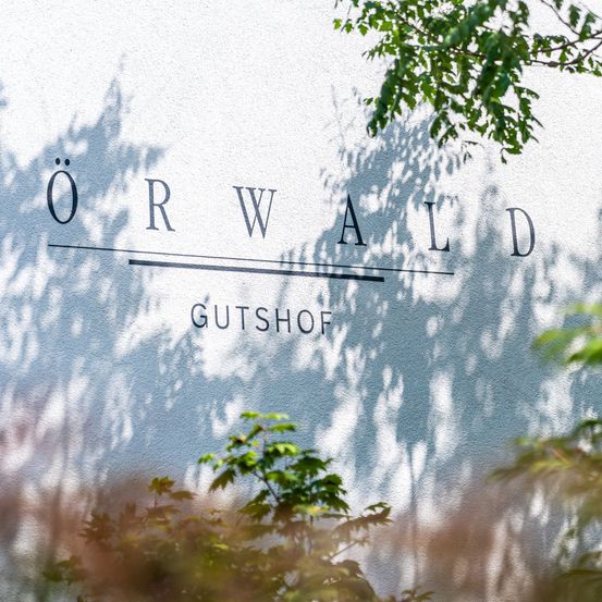 Eine weiße Wand mit schwarzem Text 'ORWALD GUTSHOF', mit Schatten von Bäumen darauf.