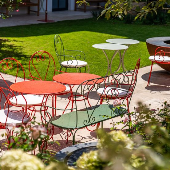 Ein Patio mit mehreren roten und grünen Stühlen und Tischen, umgeben von grünem Gras und Pflanzen, mit einer Holzbank im Hintergrund.