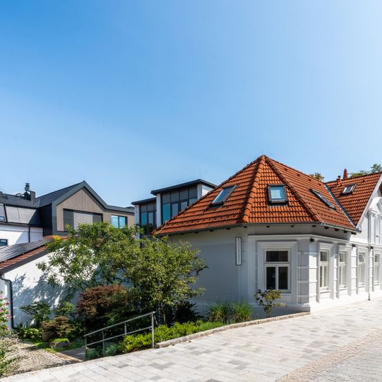 Ein modernes weißes Haus mit einem roten Dach und mehreren Fenstern, umgeben von Bäumen und Pflanzen.