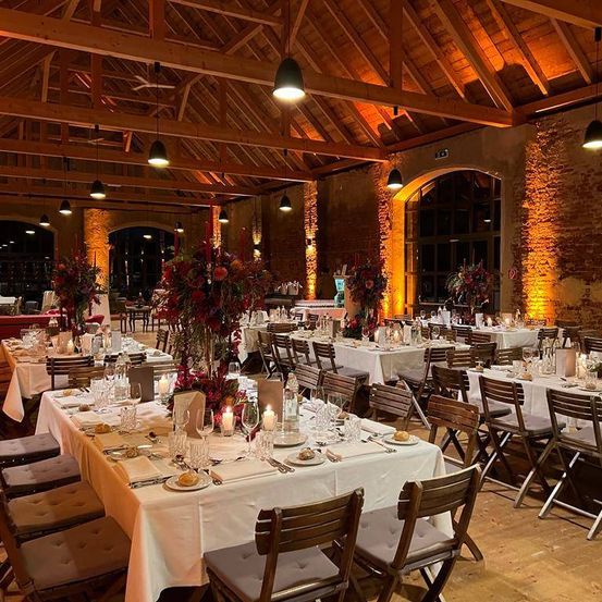 Ein Restaurant mit Holzdecke und Ziegelwänden, geschmückt mit warmer Beleuchtung. Tische sind mit Blumen, Kerzen, Tellern und Gläsern gedeckt. Die Stühle sind aus Holz, und der Boden ist aus Holz.