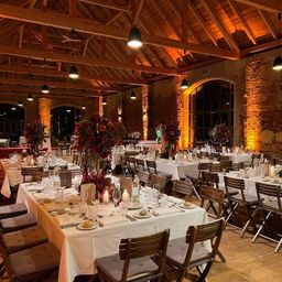 A restaurant with a wooden ceiling and brick walls, adorned with warm lighting. Tables are set with flowers, candles, plates, and glasses. The chairs are wooden, and the floor is wooden.