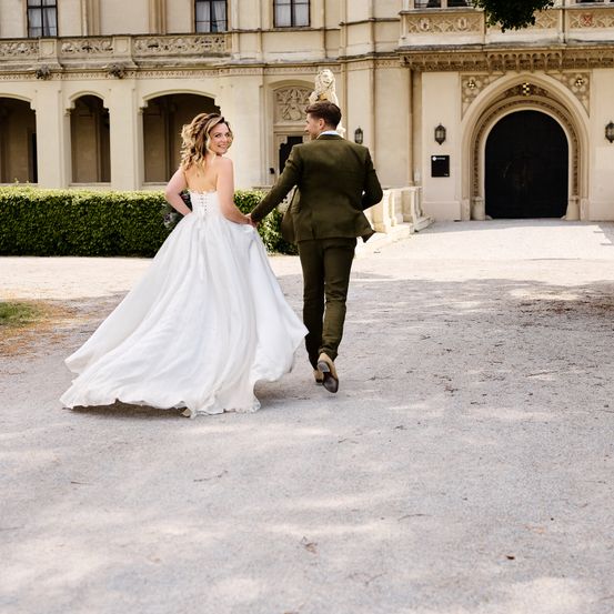 Eine Braut und ein Bräutigam gehen Hand in Hand vor einem klassizistischen Gebäude mit kunstvollen Details und einem großen Bogeneingang.