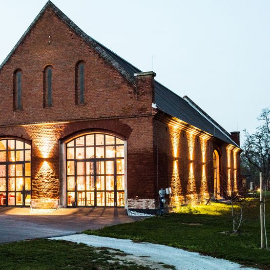 Beleuchtetes Backsteingebäude mit großen Fenstern und Rundbogentüren bei Nacht. Der Eingang wird von zwei Säulen mit Lichtern flankiert.