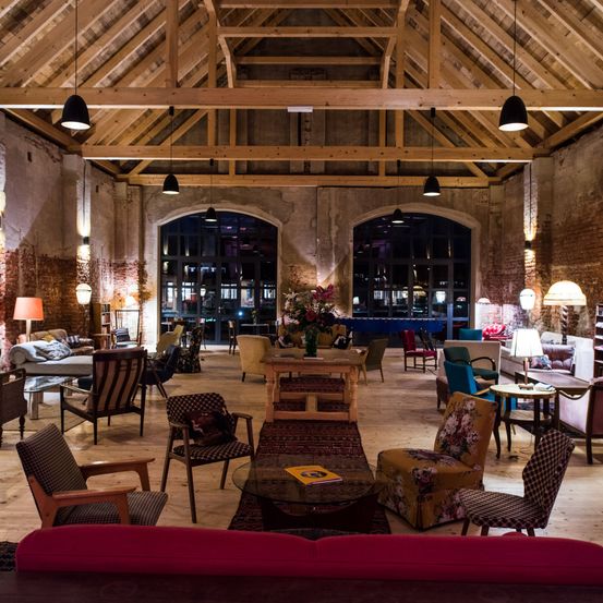 Ein gemütlicher Innenbereich mit Holzbalken und Pendelleuchten. Es gibt eine Vielzahl von Stühlen, Tischen und Lampen. Es gibt einen Teppich, Blumen und ein Buch auf einem Tisch. Es gibt auch Sofas, ein Regal und Fenster.