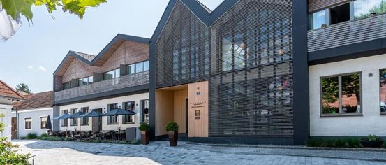 A modern architectural building with a triangular roof, a weather vane, and a wooden door. It has a glass facade and a balcony with umbrellas.