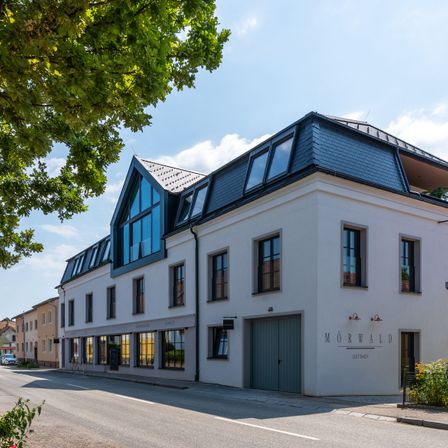 A white building with a blue roof and windows is located on a street. The building has the name Morwald on the front. There is a tree on the left side of the building.