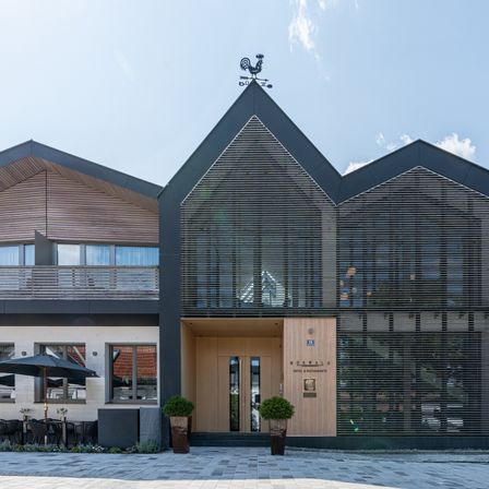 The exterior of a modern hotel building, featuring a weather vane on top, wooden and glass elements, and an entrance with two doors.
