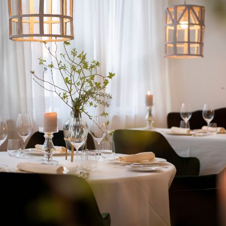 A restaurant setting with two tables, one with a plant, a candle, and wine glasses. Another table has a folded napkin, a wine glass, and a candle. White curtains and lamps hang above.