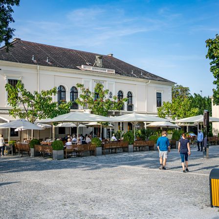 A vibrant outdoor dining area with multiple umbrellas and tables, near a historic building under a clear sky. People walk around, enjoying the sunny day.