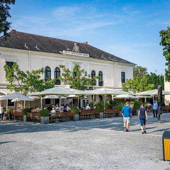 A vibrant outdoor dining area with multiple umbrellas and tables, near a historic building under a clear sky. People walk around, enjoying the sunny day.