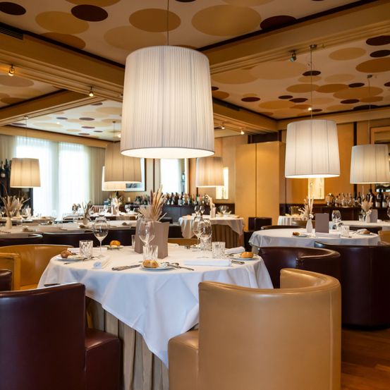 An upscale restaurant with several tables, each set with plates, glasses, and utensils. Large lamps hang from the ceiling, and the room has a modern and elegant decor.